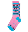 Shown stacked flat, a pair of unisex cotton crew Gumball Poodle brand sock in pink with black and teal stripes around the leg and a teal heel and toe. The text on the sock reads, "BE THE FLAMINO IN A FLOCK OF PIDGEONS".