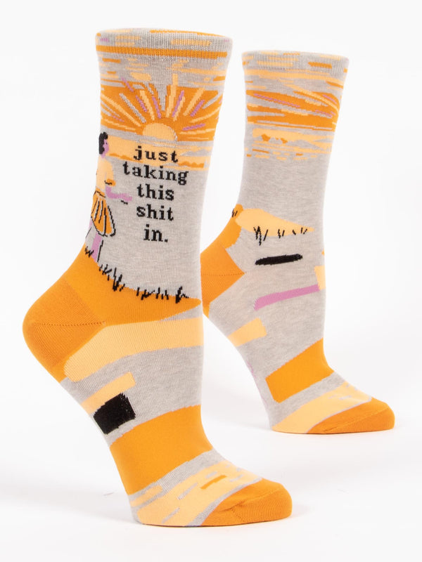 Shown on leg forms, a pair of light grey socks with an orange heel and toe. These socks feature a cartoon woman watching the sunset with the phrase, "I'm just taking this shit in." on the leg.