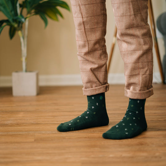 a person standing on a wood floor wearing beige plaid pants and green conscious step socks with a design of small trees all over them