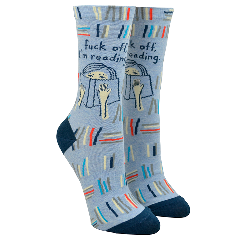 Shown on leg forms with the text side out, a pair of Blue Q brand combed cotton women's crew sock in denim blue with a navy blue heel and toe. The sock features an all over design of abstracted stacks of library books in shades of blue, grey, and red. The leg of the sock has two designs, one side has an illustration of a sleeping cat and the other has a cartoon women reading a book with the words 