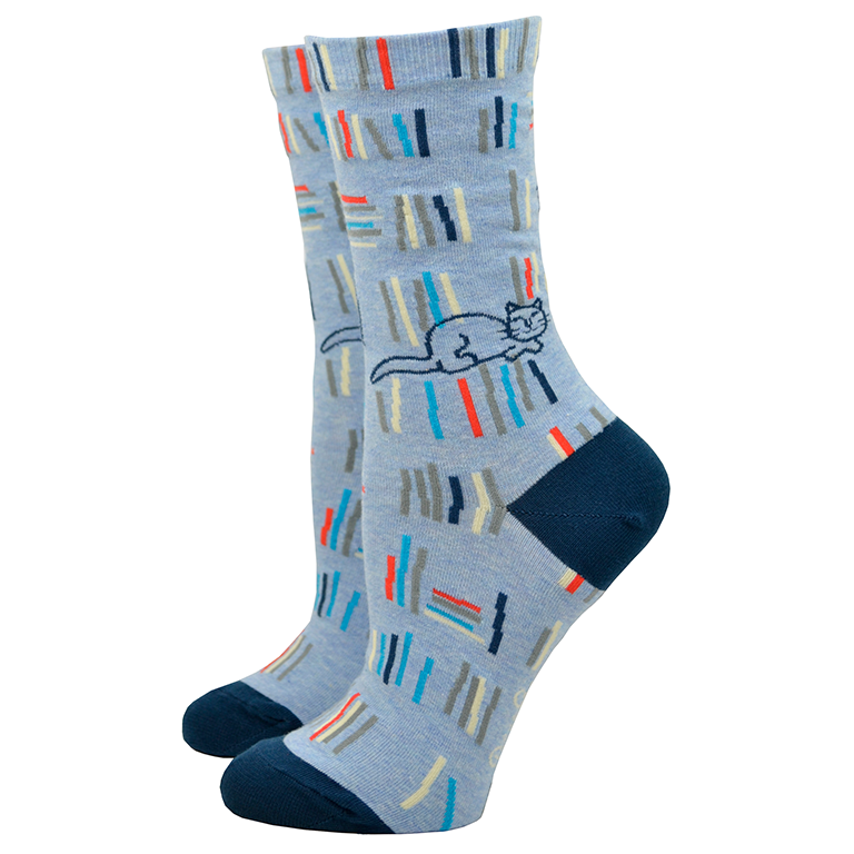 Shown on leg forms with the cat side out, a pair of Blue Q brand combed cotton women's crew sock in denim blue with a navy blue heel and toe. The sock features an all over design of abstracted stacks of library books in shades of blue, grey, and red. The leg of the sock has two designs, one side has an illustration of a sleeping cat and the other has a cartoon women reading a book with the words 