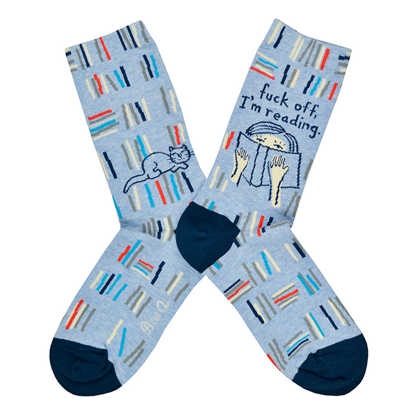 Shown in a flatlay, a pair of Blue Q brand combed cotton women's crew sock in denim blue with a navy blue heel and toe. The sock features an all over design of abstracted stacks of library books in shades of blue, grey, and red. The leg of the sock has two designs, one side has an illustration of a sleeping cat and the other has a cartoon women reading a book with the words " fuck off, I'm reading." above her.