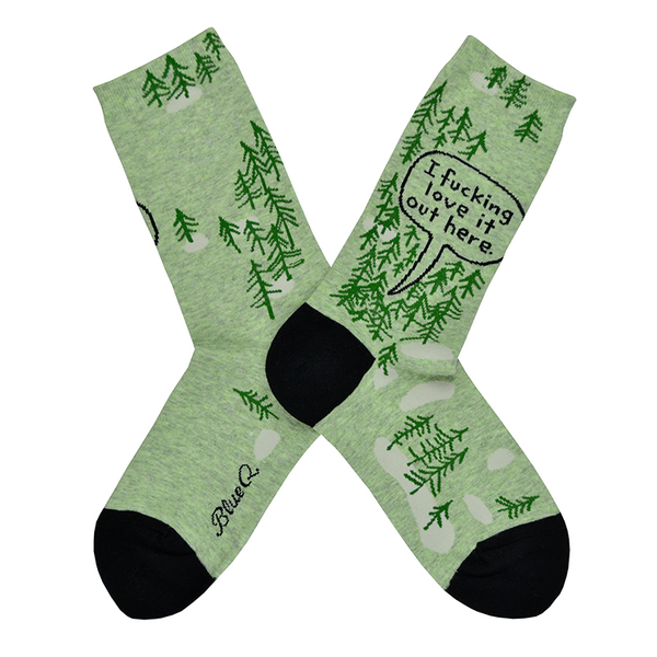 Shown in a flatlay, a pair of women's Blue Q brand combed cotton crew socks in green with a black heel and toe. The socks feature a cartoon-y pine tree motif all over the sock with the words, "I fucking love it out here." in a black speech bubble