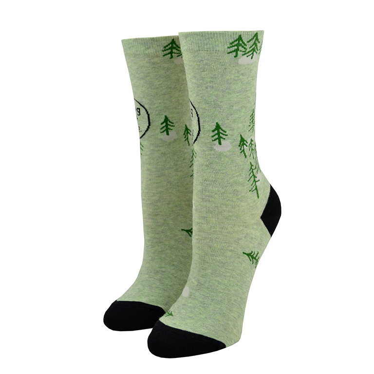 Shown on leg forms, a pair of women's Blue Q brand combed cotton crew socks in green with a black heel and toe. The socks feature a cartoon-y pine tree motif all over the sock with the words, 