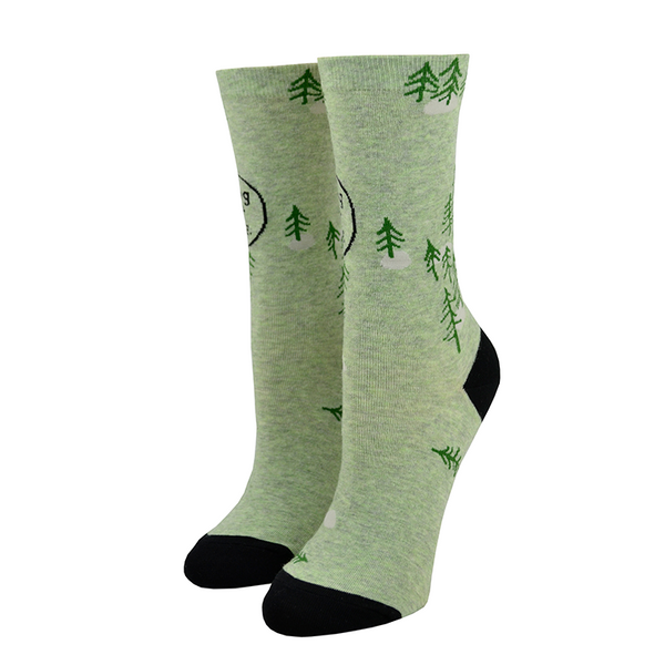 Shown on leg forms, a pair of women's Blue Q brand combed cotton crew socks in green with a black heel and toe. The socks feature a cartoon-y pine tree motif all over the sock with the words, "I fucking love it out here." in a black speech bubble.