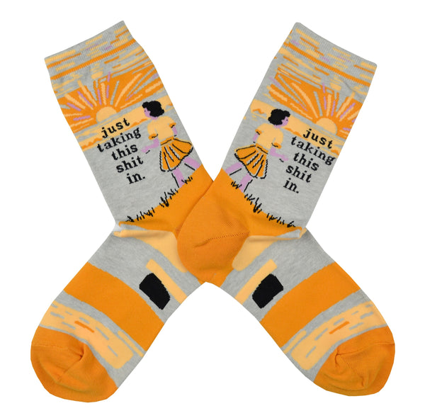 Shown in a flatlay, a pair of light grey socks with an orange heel and toe. These socks feature a cartoon woman watching the sunset with the phrase, "I'm just taking this shit in." on the leg.
