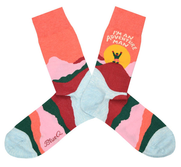 Shown in a flatlay, a pair of men's Blue Q brand combed cotton crew socks with a peach cuff and light blue heel and toe. This sock features an abstract mountain design in shades pink, blue, and green with a cartoon man in front of the sun on the leg. The socks reads, "I'm an adventure man!" on the leg.