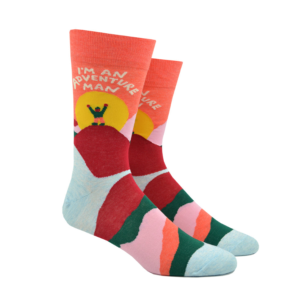 Shown on leg forms, a pair of men's Blue Q brand combed cotton crew socks with a peach cuff and light blue heel and toe. This sock features an abstract mountain design in shades pink, blue, and green with a cartoon man in front of the sun on the leg. The socks reads, 