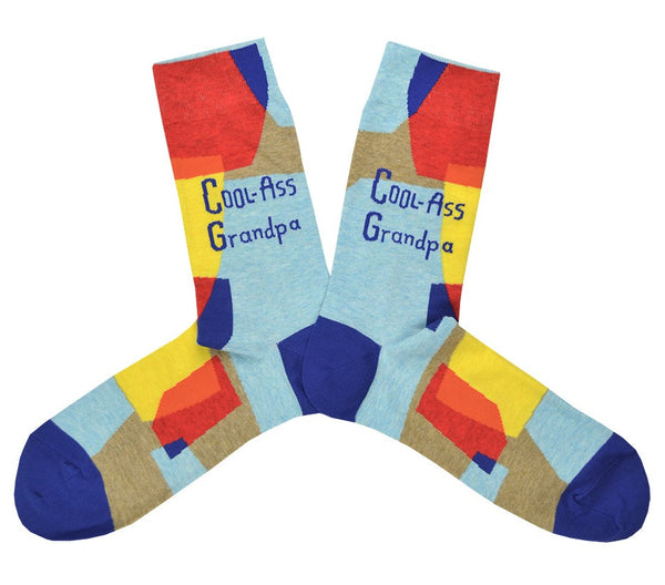 Shown in a flatlay, a pair of Blue Q cotton men’s crew socks with geometric blue, red and yellow shapes, plus the words “Cool Ass Grandpa”