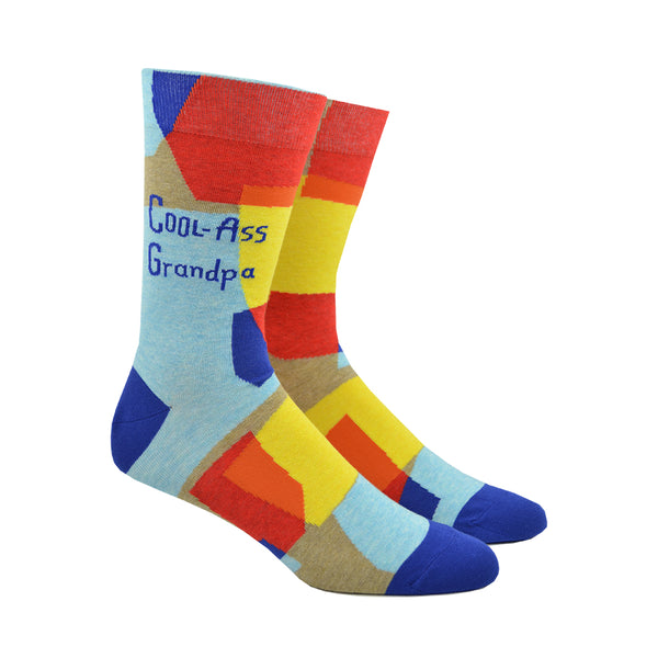 Shown on a leg form, a pair of Blue Q cotton men’s crew socks with geometric blue, red and yellow shapes, plus the words “Cool Ass Grandpa”