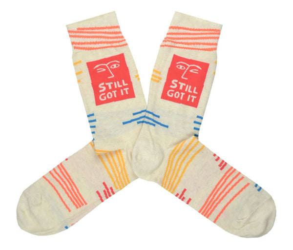 Shown in a flatlay, a pair of Blue Q cream-white cotton men’s crew socks with orange abstract winking face, “Still Got It” text and abstract accent lines in blue, orange, and yellow
