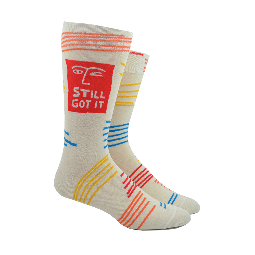 Shown on a leg form, a pair of Blue Q cream-white cotton men’s crew socks with orange abstract winking face, “Still Got It” text and abstract accent lines in blue, orange, and yellow