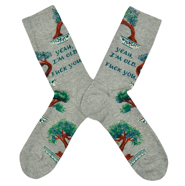 Shown in a flatlay, a pair of Blue Q brand men's cotton crew sock in grey with bonsai trees scattered around the sock. The text on the leg of the sock reads, "Yeah, I'm Old. Fuck You.".