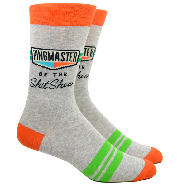 Shown on a leg form, a pair of Blue Q gray cotton men’s crew socks with orange cuff/heel/toe, green accent tripe and the words “Ringmaster of the ShitShow”
