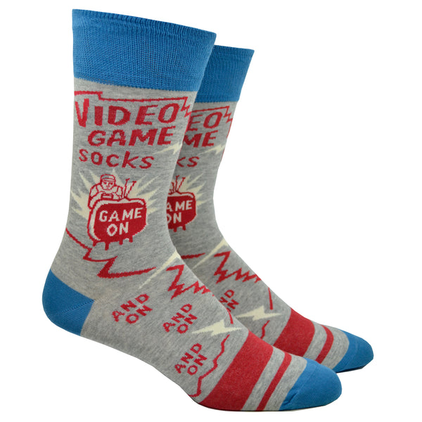 Shown on leg forms, a pair of Blue Q brand men's cotton crew socks in grey with a blue cuff/heel/toe and 2 red stripes on the foot. The socks feature a cartoon man gaming in front of a TV with text that starts at the top of the sock and goes down onto the foot reading, "Video Game Socks, Game On, and On and On and On".