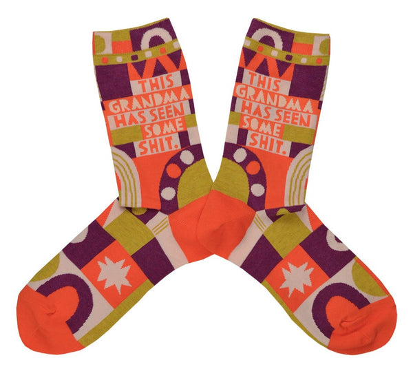 A pair of brightly multi-colored womens socks with the text "This Grandma Had Seen Some Shit." in orange and white. The socks feature abstract shapes and design elements in orange, white, gold and, purple. 