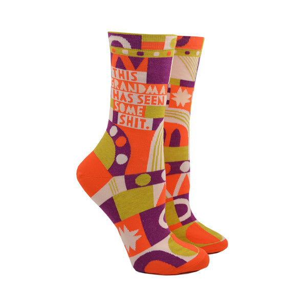 Shown on leg forms, a pair of brightly multi-colored womens socks with the text "This Grandma Had Seen Some Shit." in orange and white. The socks feature abstract shapes and design elements in orange, white, gold and, purple.