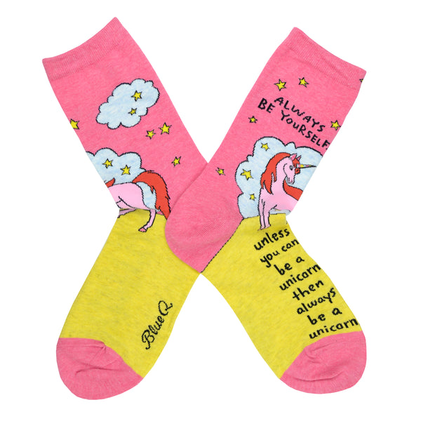 These pink cotton women's crew socks by the brand Blue Q feature a unicorn standing on a yellow hill in front of a blue cloud and yellow stars with the quote "Always be yourself" on the leg, continuing "unless you can be a unicorn, then always be a unicorn" written on the foot.