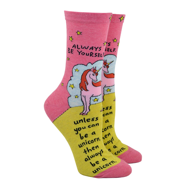 Shown on a leg form, these pink cotton women's crew socks by the brand Blue Q feature a unicorn standing on a yellow hill in front of a blue cloud and yellow stars with the quote "Always be yourself" on the leg, continuing "unless you can be a unicorn, then always be a unicorn" written on the foot.