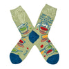 Shown in a flatlay, a pair of women's Blue Q cotton crew socks in olive green with a blue heel and toe. These socks feature a cartoon women over a pot with a spoon and the phrase, "Get the hell out of My kitchen" above her. The foot of the sock features abstracted pots and pans in red, green, and orange.