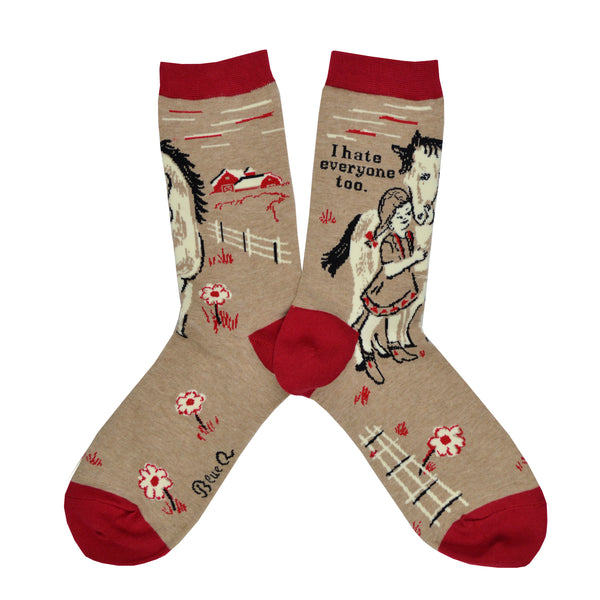 Shown in a flatlay, a pair of women's Blue Q brand combed cotton socks in brown with a red heel, toe, and cuff. The leg of the sock features a little girl with a horse with the phrase, "I hate everyone too." while the foot has a fence and flower design.