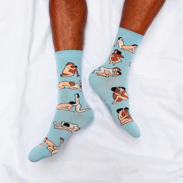 Shown on a dark skinned models feet, a pair of women's CouCou Suzette cotton crew socks in light blue with gay male lovers engaging in varying Kama Sutra sex positions all over the socks.