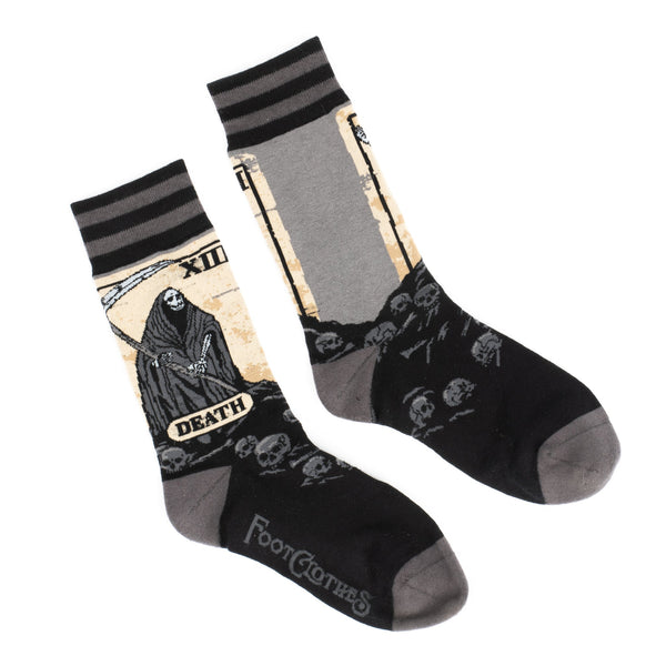 Shown in a flatlay, a pair of unisex crew sock in black and grey. The leg of the sock features a depiction of the Death tarot card with the Grim Reaper and his scythe. The foot of the sock is black with grey skulls and bones descending below the reaper.