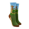 Shown on leg forms, a pair of women's Foot Traffic brand cotton crew socks with a blue cuff and brown heel and toe. The leg of the sock has a giraffe head and neck with a savanna background. The foot is dark green on the top and the sole is a giraffe pattern.