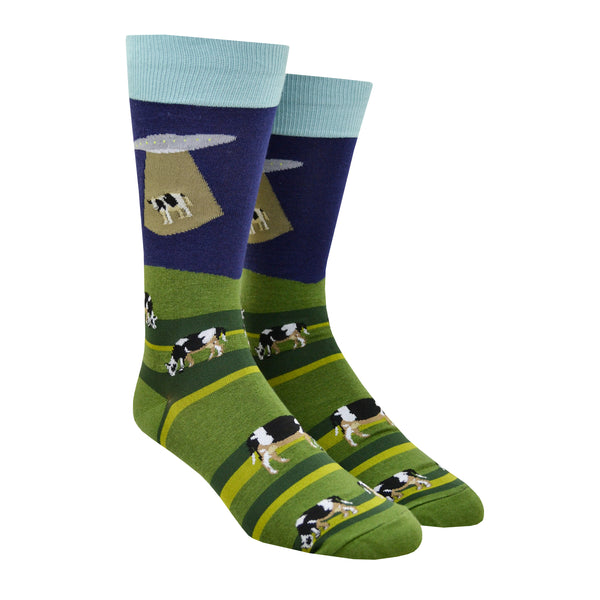 Shown on a leg form, these blue and green cotton men's crew socks with a light blue cuff by the brand Foot Traffic feature an alien spaceship beaming up a cow that was grazing in a field.