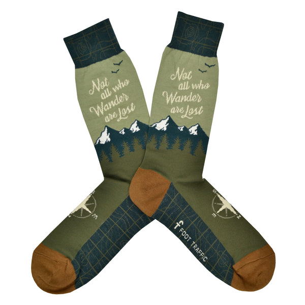 Shown in a flatlay, a pair of men's Foot Traffic brand cotton crew socks with a dark blue cuff, green heel, and mustard toe. The leg of this sock is light green with a blue mountain rage and the words, "Not all who wander are lost" in a cursive font. The foot is green with a white compass on the top of the foot and a dark blue map on the bottom of the foot.