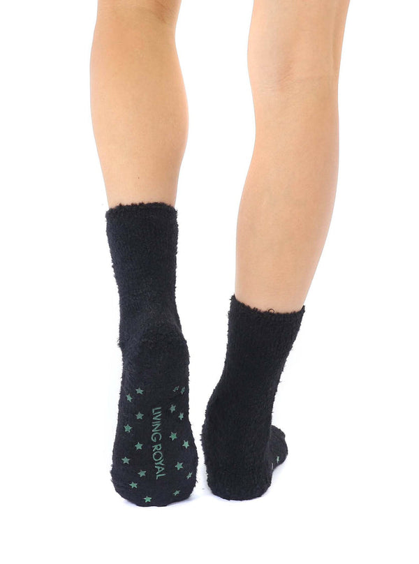A pair of black fuzzy socks with an embroidered green alien face on the front of the calf shown from the back.