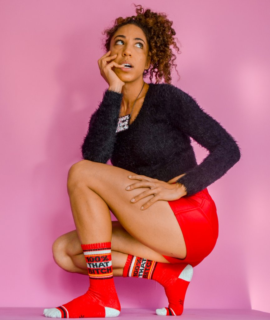 Shown on a dark skinned feminine model crouched in front of a pink backdrop, a pair of cotton crew length unisex Gumball Poodle brand socks in red with a sparkly silver toe and heel. The leg of the sock features, black, sparkly silver, and yellow stripes with the phrase, 