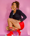 Shown on a dark skinned feminine model crouched in front of a pink backdrop, a pair of cotton crew length unisex Gumball Poodle brand socks in red with a sparkly silver toe and heel. The leg of the sock features, black, sparkly silver, and yellow stripes with the phrase, "100% That Bitch" along the cuff.
