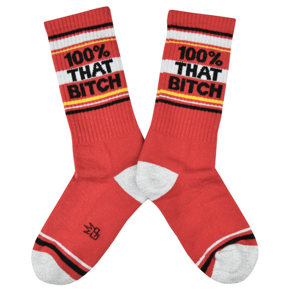 Shown in a flatlay, a pair of cotton crew length unisex Gumball Poodle brand socks in red with a sparkly silver toe and heel. The leg of the sock features, black, sparkly silver, and yellow stripes with the phrase, "100% That Bitch" along the cuff.