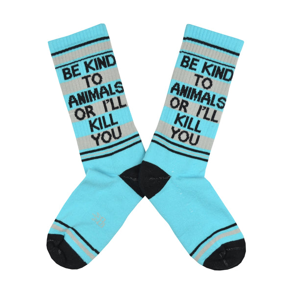 Shown in a flatlay, a pair of unisex cotton crew Gumball Poodle brand socks in light blue with black, blue, and grey stripes around the leg and cuff. The heel and toe are black and the leg of the sock reads in black font, BE KIND TO ANIMALS OR I'LL KILL YOU".