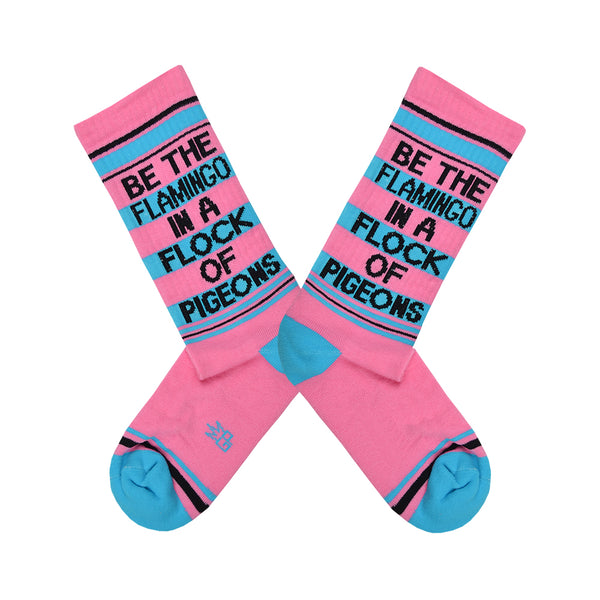 Shown in a flatlay, a pair of unisex cotton crew Gumball Poodle brand sock in pink with black and teal stripes around the leg and a teal heel and toe. The text on the sock reads, "BE THE FLAMINO IN A FLOCK OF PIDGEONS".