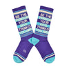 Shown in a flatlay, a pair of Gumball Poodle brand unisex cotton crew sock in purple with cream and blue stripes around the leg and a blue heel and toe. On the side of the sock in cream lettering it says, "BE THE PERSON YOUR DOG THINKS YOU ARE".