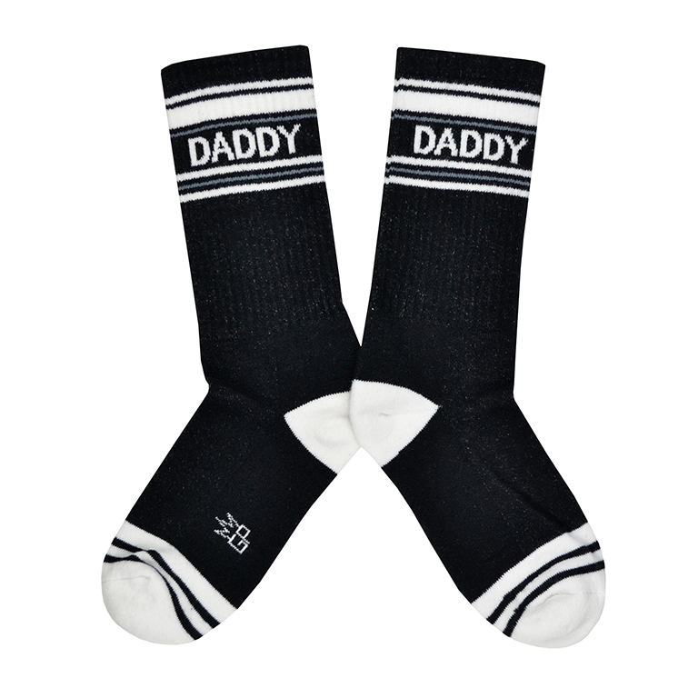 Shown in a flatlay, a pair of Gumball Poodle, black cotton crew socks with white heel/toe/accent stripes and “Daddy” text