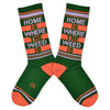 Shown in a flatlay, a pair of Gumball Poodle unisex cotton crew sock in green with an orange and green striped toe and cuff. The leg features the words, "HOME IS WHERE THE WEED IS".