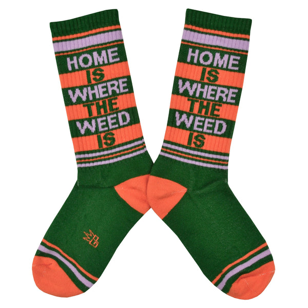 Shown in a flatlay, a pair of Gumball Poodle unisex cotton crew sock in green with an orange and green striped toe and cuff. The leg features the words, "HOME IS WHERE THE WEED IS".