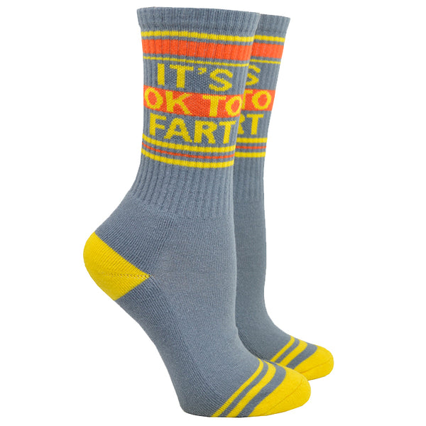 Shown on leg forms, a pair of grey cotton Gumball Poodle brand unisex crew socks with yellow and orange stripes on the cuff and yellow striped heel and toe. These socks feature the phrase, "IT'S OKAY TO FART" in yellow type on the leg.