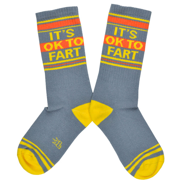 Shown in a flatlay, a pair of grey cotton Gumball Poodle brand unisex crew socks with yellow and orange stripes on the cuff and yellow striped heel and toe. These socks feature the phrase, "IT'S OKAY TO FART" in yellow type on the leg.
