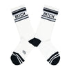 These white cotton unisex crew socks with a black toe and striped cuff by the brand Gumball Poodle feature the words "BITCH" on the leg.