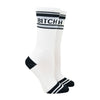 Shown on a leg form, these white cotton unisex crew socks with a black toe and striped cuff by the brand Gumball Poodle feature the words "BITCH" on the leg.