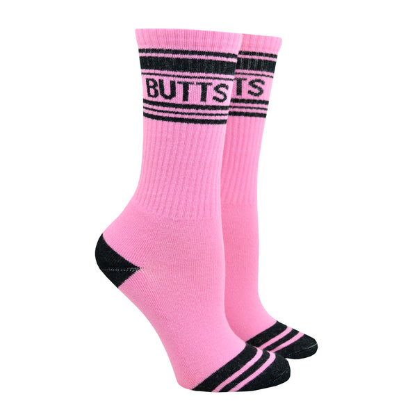 Shown on a leg form, these pink cotton unisex crew socks with a black striped toe and cuff by the brand Gumball Poodle feature the word "BUTTS" on the leg.