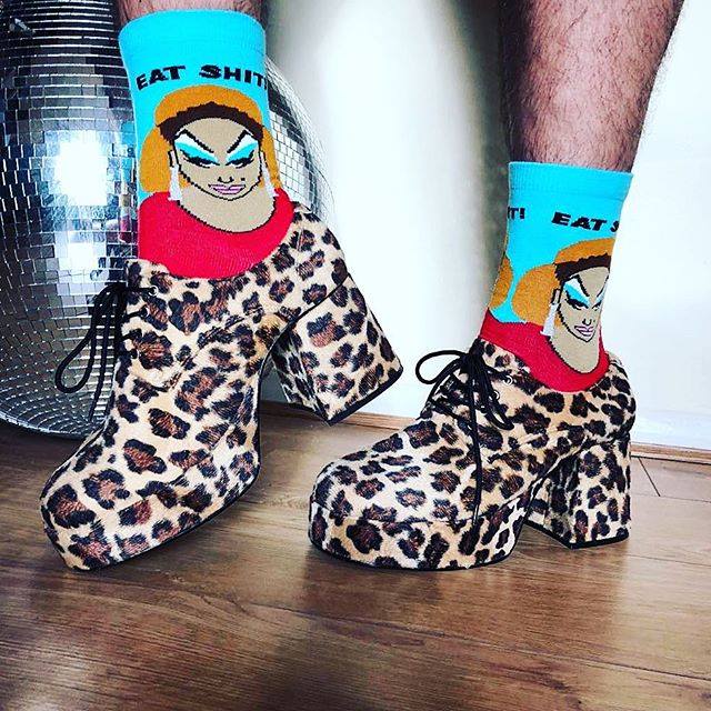A model wearing blue and red cotton unisex crew socks feature a portrait of the legendary drag queen Divine and the words 