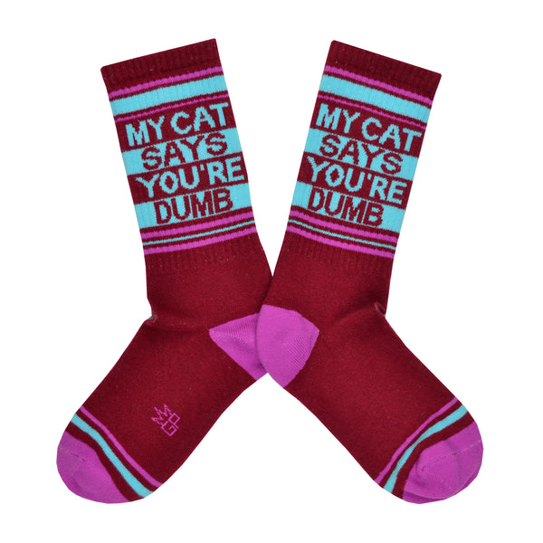 Shown in a flatlay, a pair of Gumball Poodle brand unisex cotton crew socks in maroon with a magenta heel/toe and a blue striped cuff that says, 'MY CAT SAYS YOUR'RE DUMB" on the leg.