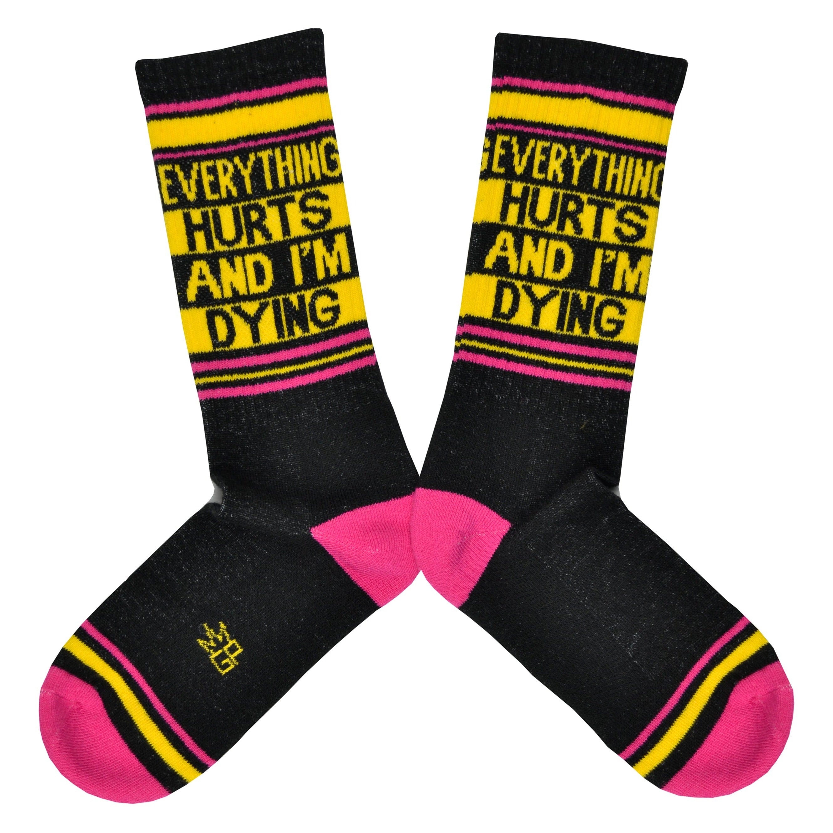 Shown in a flatlay, a pair of Gumball Poodle black cotton crew socks with pink toe/heel and yellow “Everything Hurts and I’m Dying” text