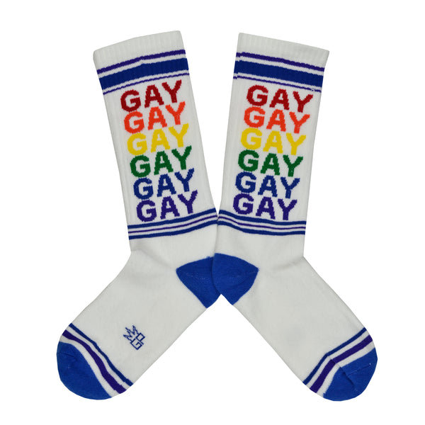 These white cotton unisex crew socks with a blue striped toe and cuff by the brand Gumball Poodle feature the word "GAY" repeated down the leg in rainbow colors.