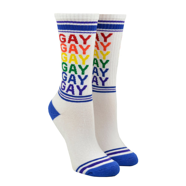Shown on a leg form, these white cotton unisex crew socks with a blue striped toe and cuff by the brand Gumball Poodle feature the word "GAY" repeated down the leg in rainbow colors.